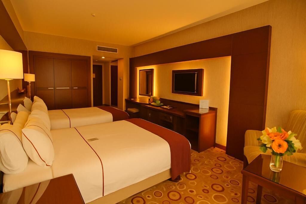 A hotel room with two beds, a desk, a chair, a TV, and a vase with flowers.