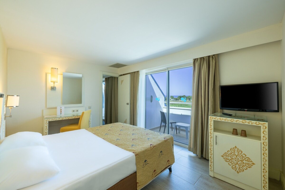 Habitación de hotel con cama, escritorio, televisor y balcón con vistas.