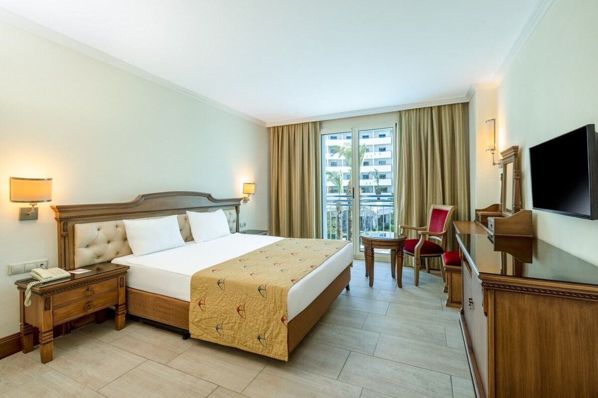 Habitación de hotel con cama, mesita de noche, lámpara, silla, escritorio, espejo, televisor y balcón con cortinas.