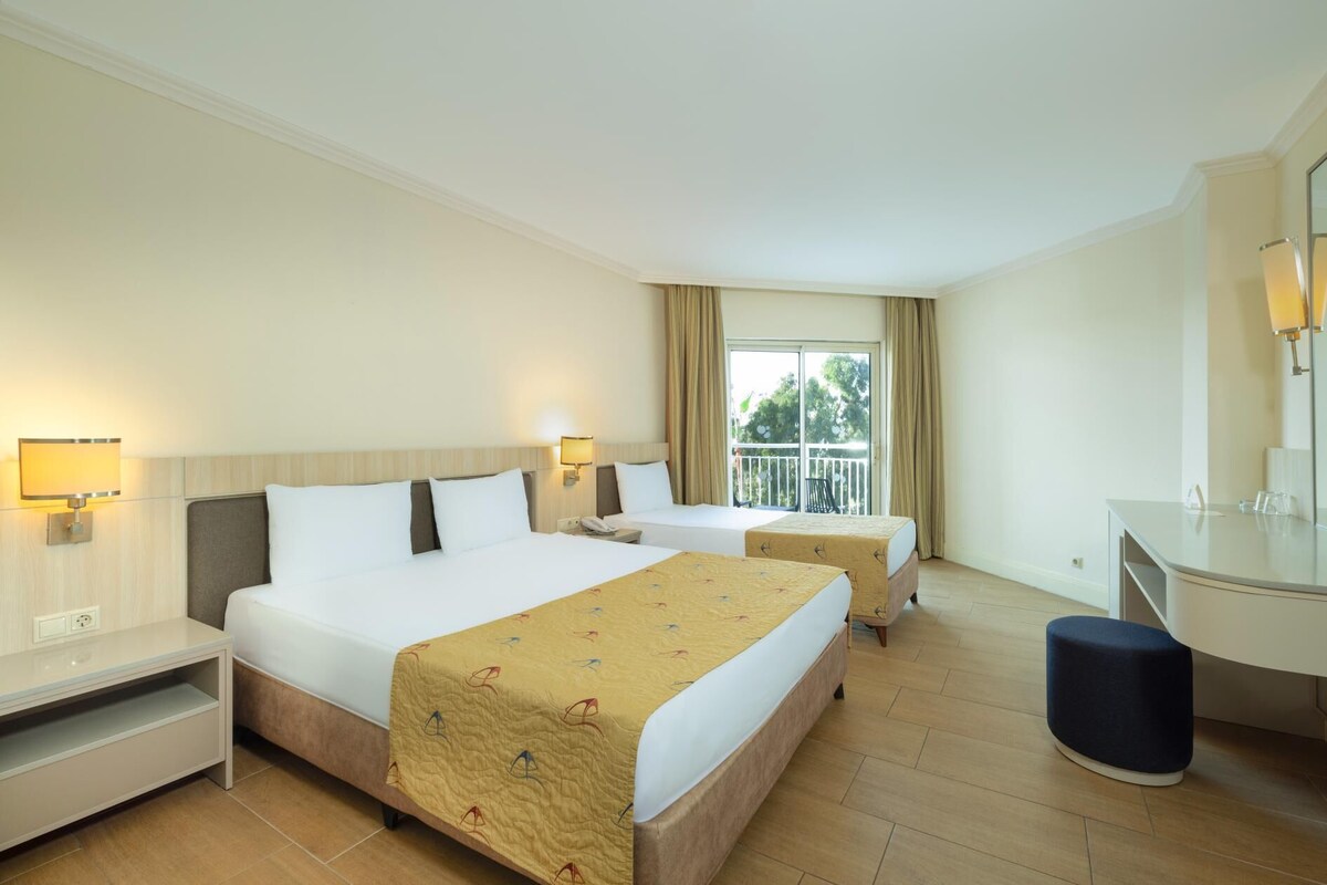 Habitación de hotel con dos camas, un escritorio, una silla, un espejo y vistas al exterior.