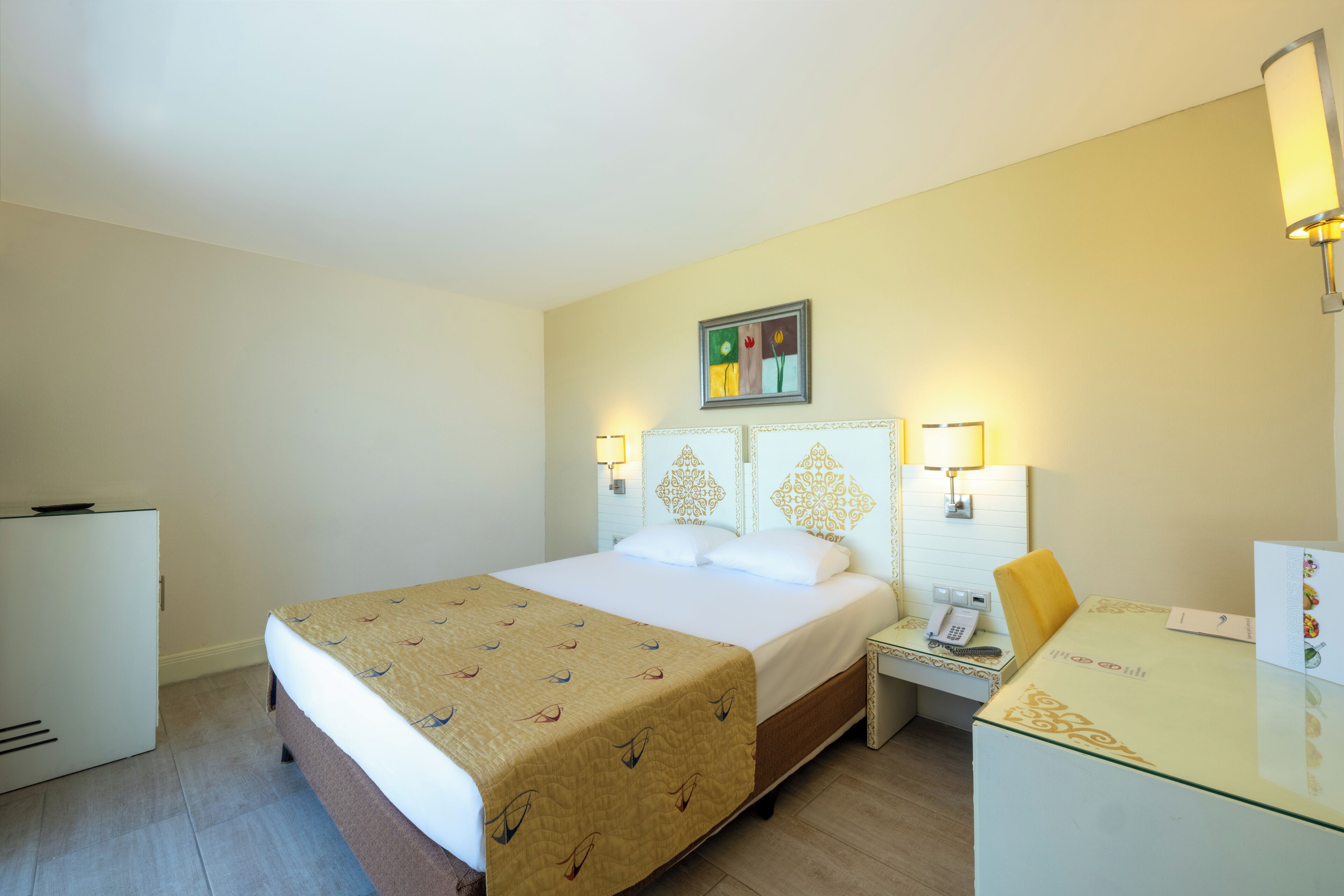 A hotel room with a bed, bedside table, lamp, desk, and a small refrigerator.