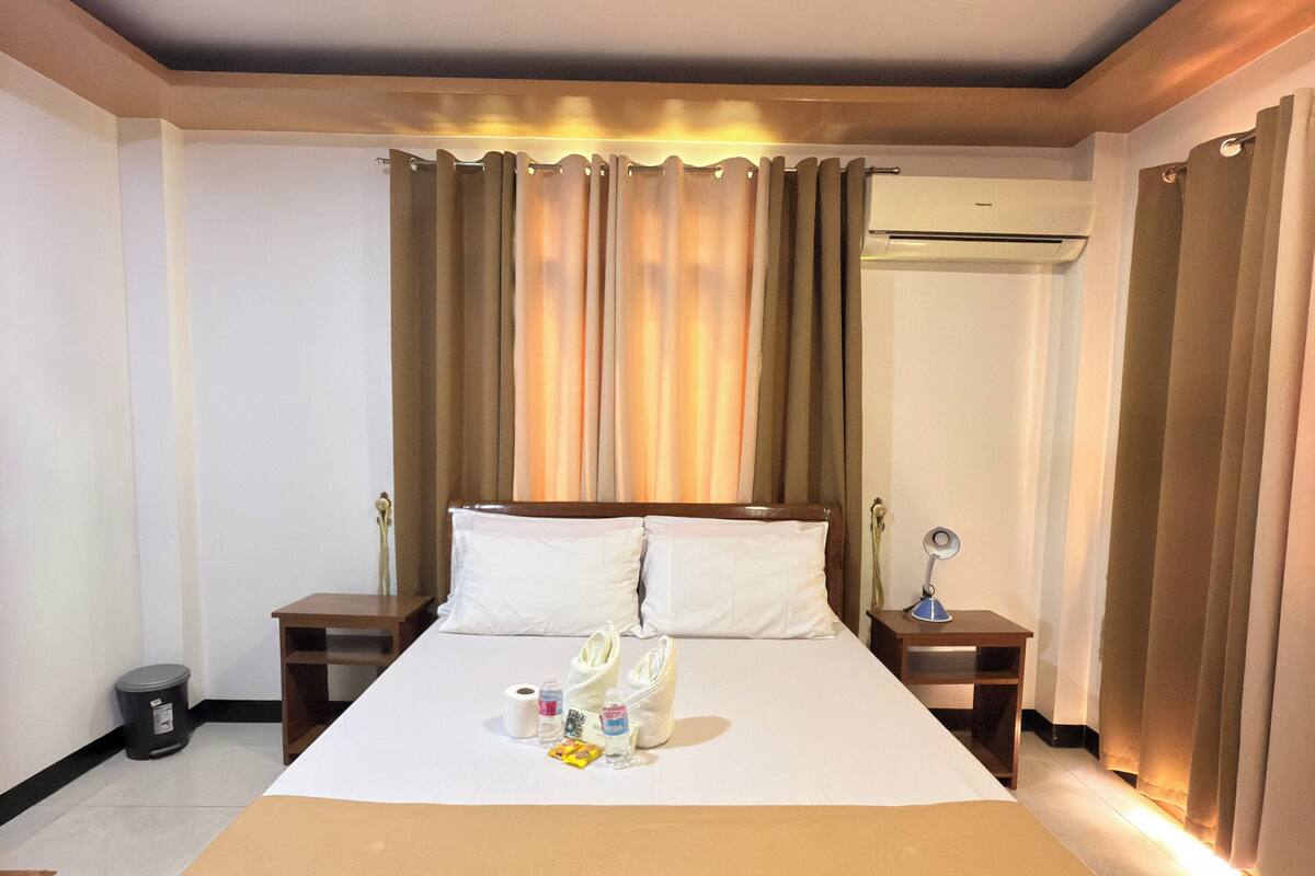 Ein Hotelzimmer mit Bett, Deckenventilator, Nachttisch mit Lampe und Vorhängen.