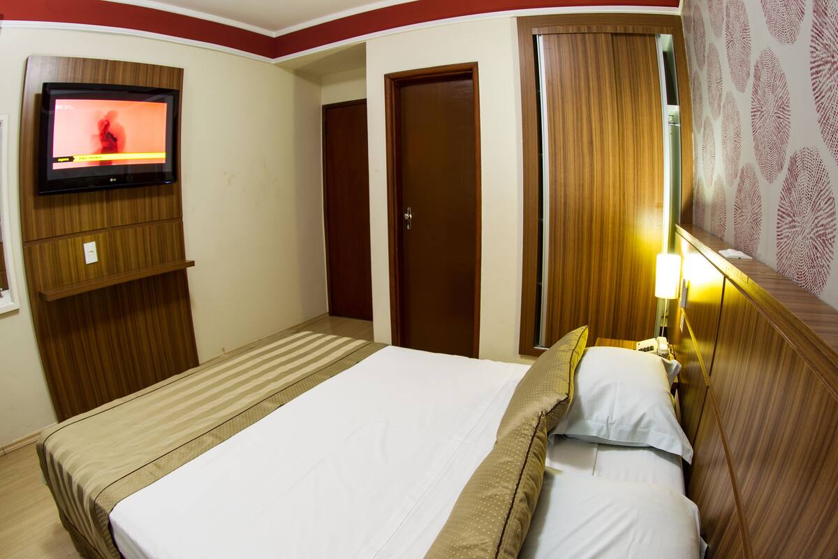 Une chambre d’hôtel avec un lit, un téléviseur fixé au mur, une armoire en bois et une porte ornée d’un rideau à motifs.