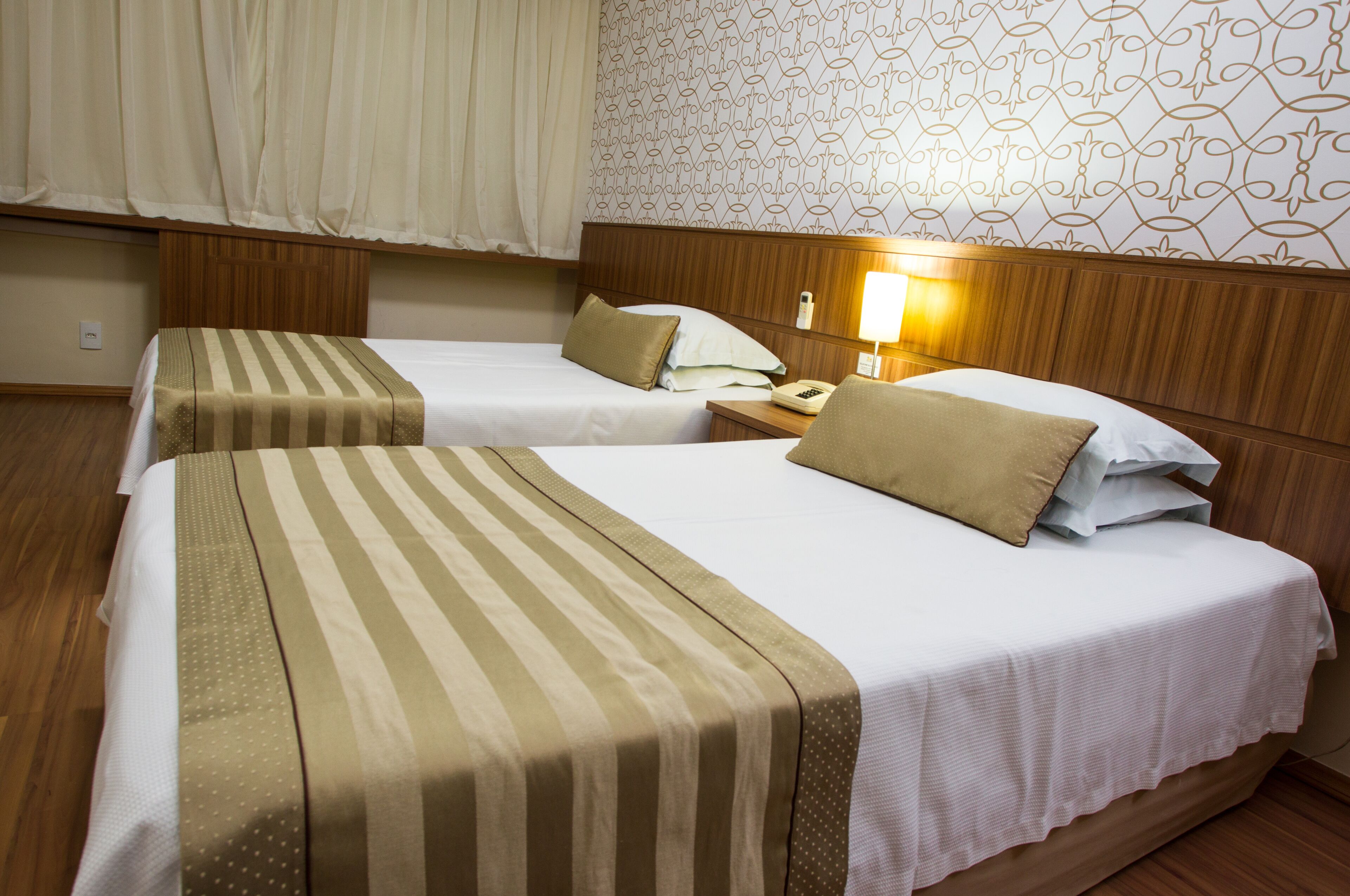 A hotel room with two beds, each with a striped bedspread and pillows. There is a nightstand with a lamp and a phone. The room has wooden paneling on the walls and a patterned wallpaper.