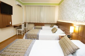 A hotel room with three beds, a TV, a desk with a lamp, and a chair.