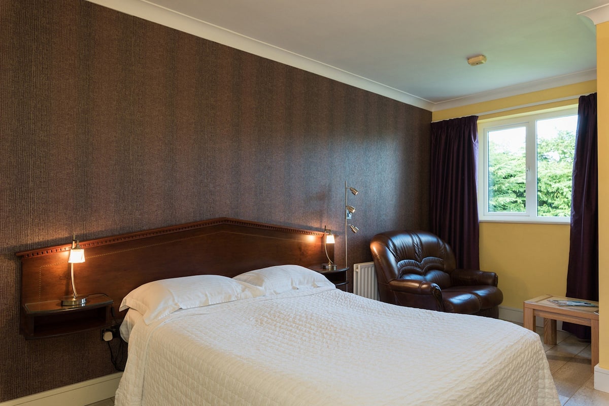 A hotel room with a large bed, a nightstand with a lamp, a leather armchair, and a window with curtains.