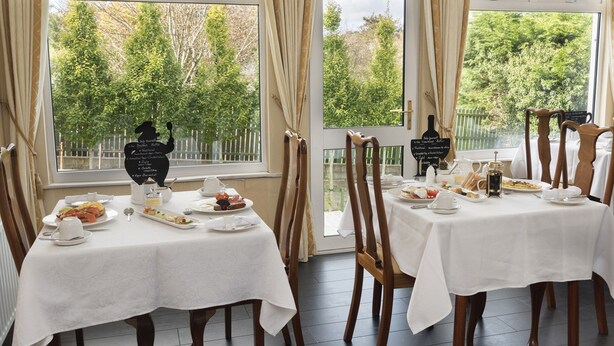 Un comedor con mesas dispuestas para el desayuno, ventanas amplias con cortinas y vistas a zonas verdes.