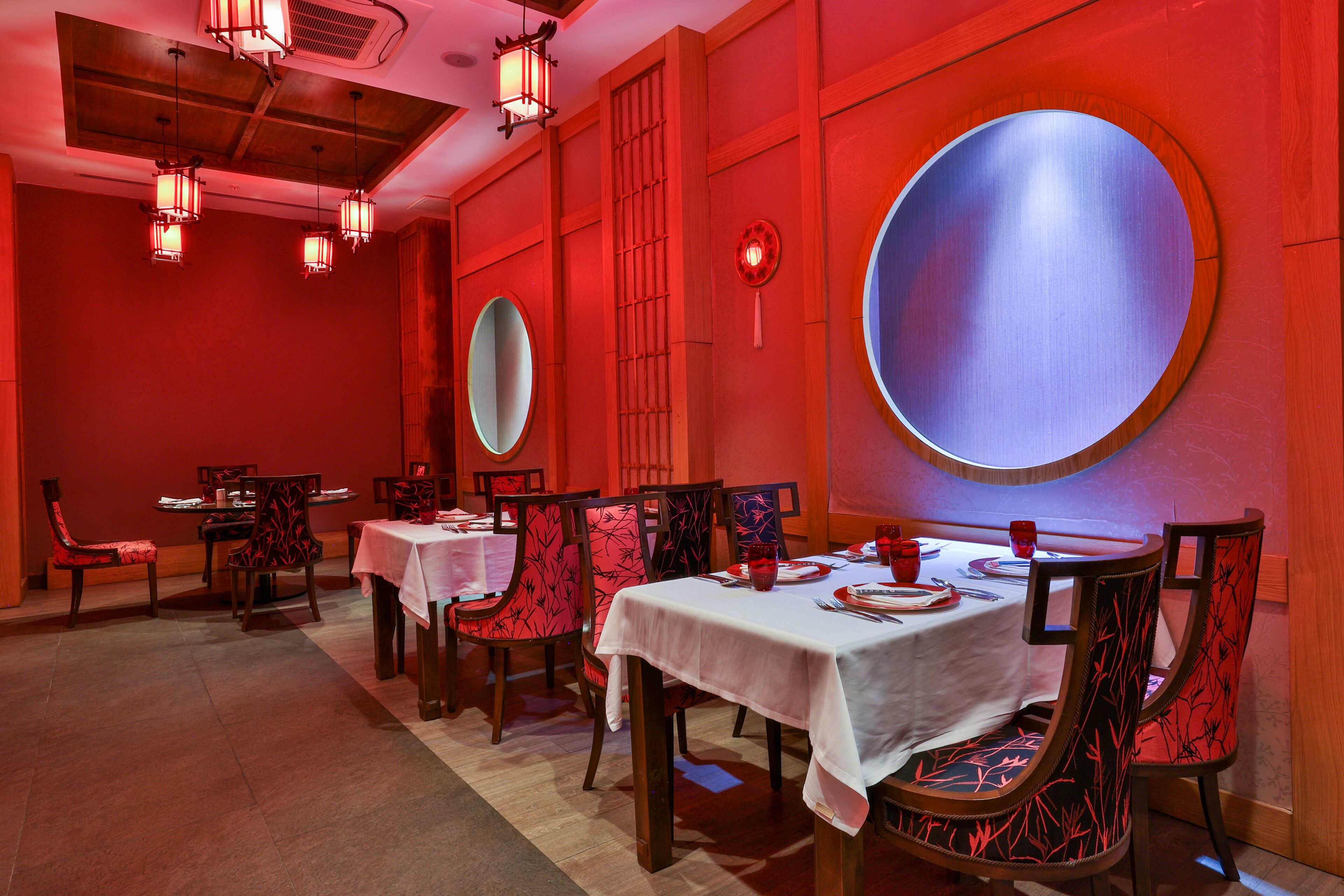 A dining area with red walls, round mirrors, and patterned chairs. Tables are set with white tablecloths and red napkins.