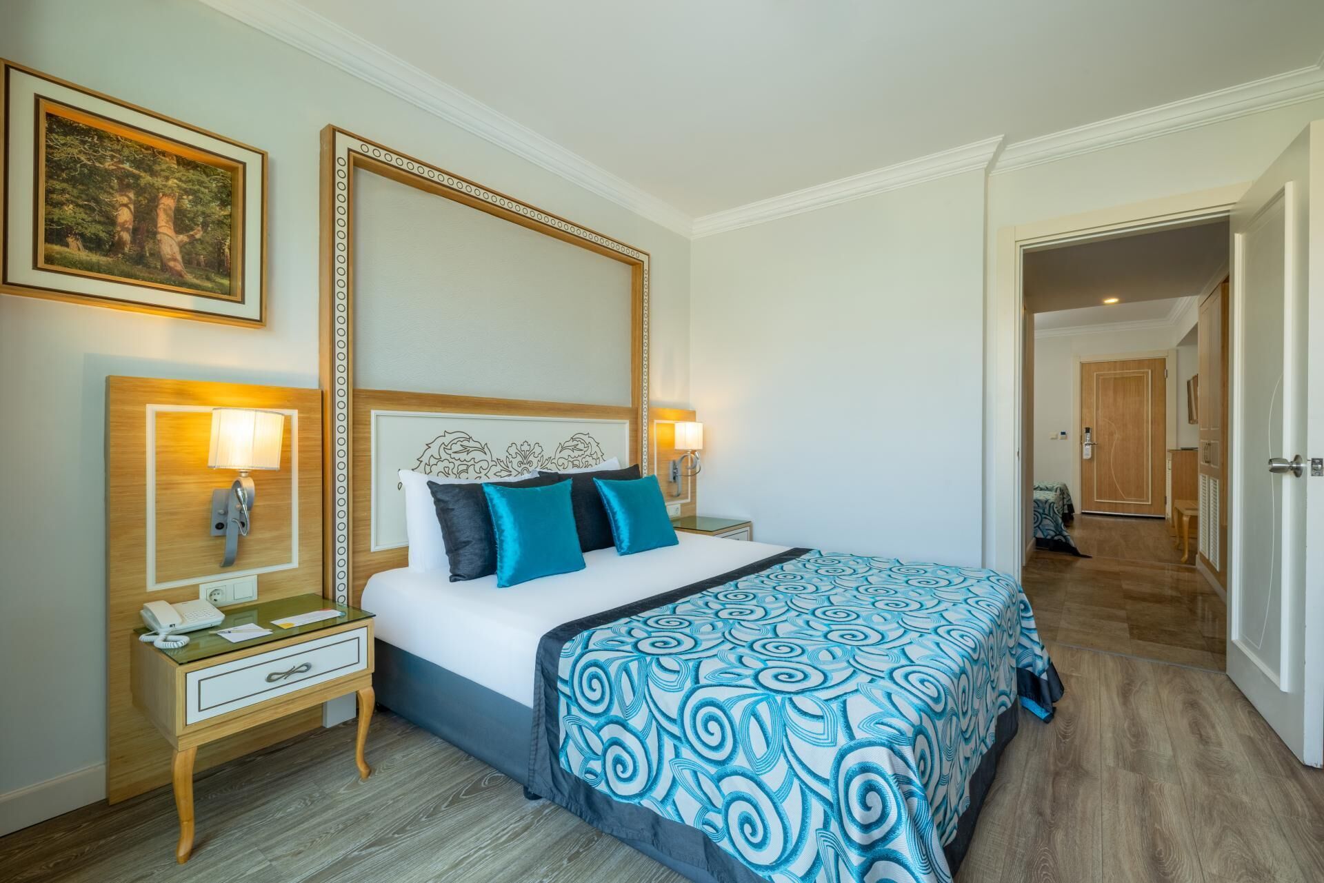 Habitación de hotel con cama, mesitas de noche, un espejo y una puerta que da a otra habitación.