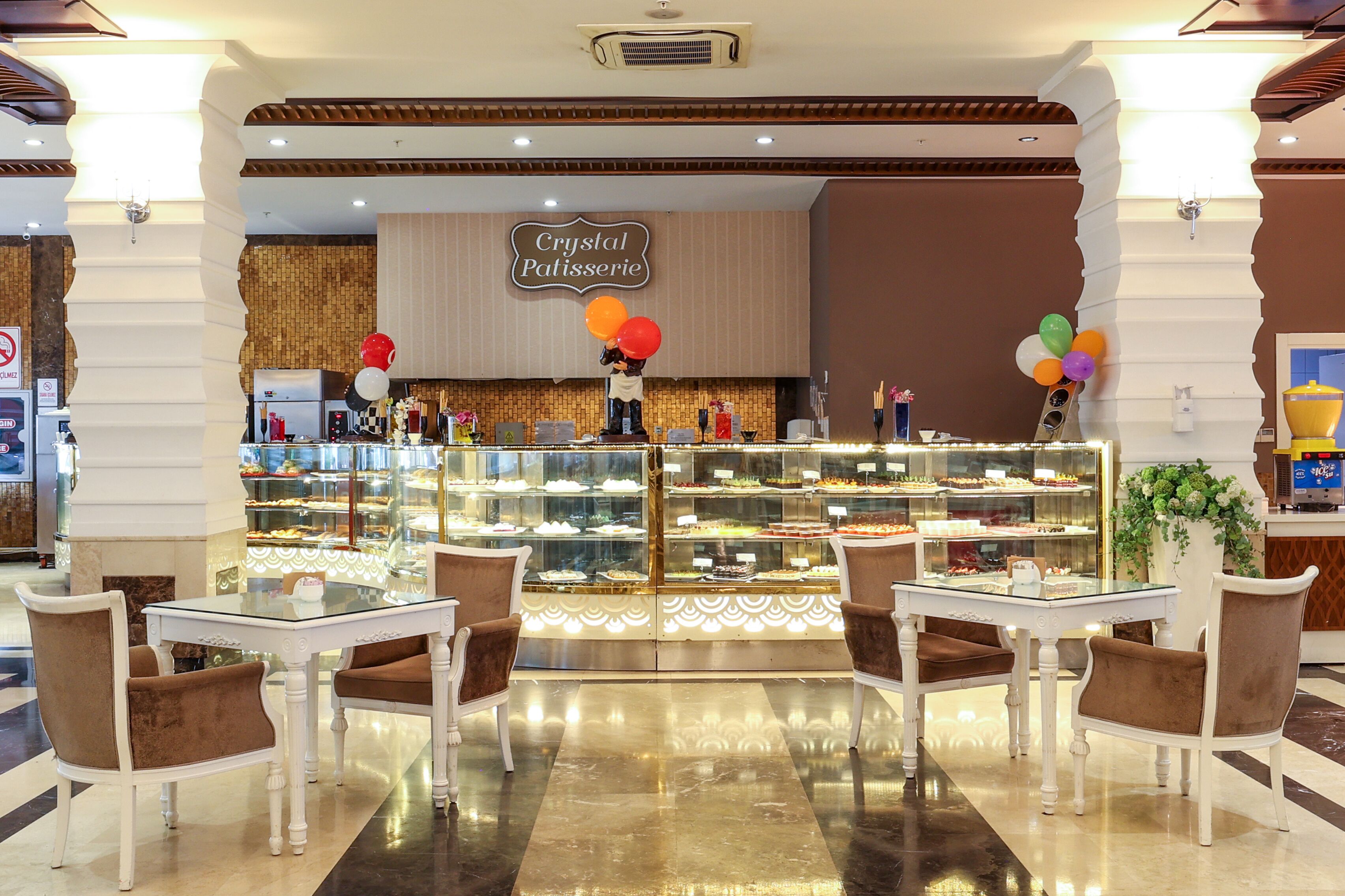 Crystal Patisserie con un expositor de vidrio lleno de pasteles, una mesa blanca con sillas marrones y globos en la barra.