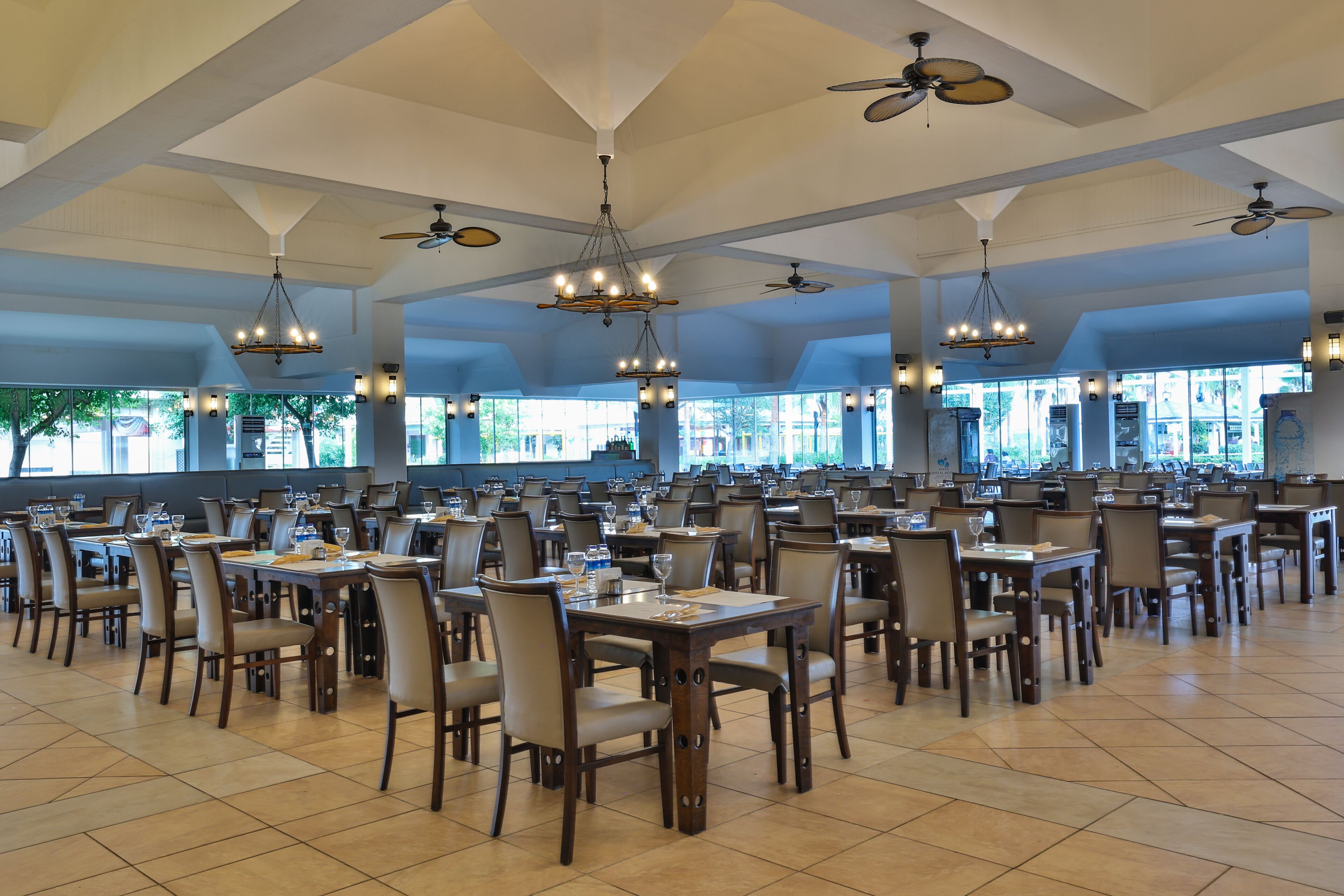 A spacious dining area with multiple tables and chairs, ceiling fans, and large windows allowing natural light.