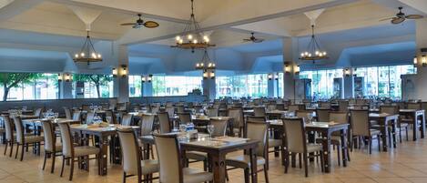 A spacious dining area with multiple tables and chairs, ceiling fans, and large windows allowing natural light.