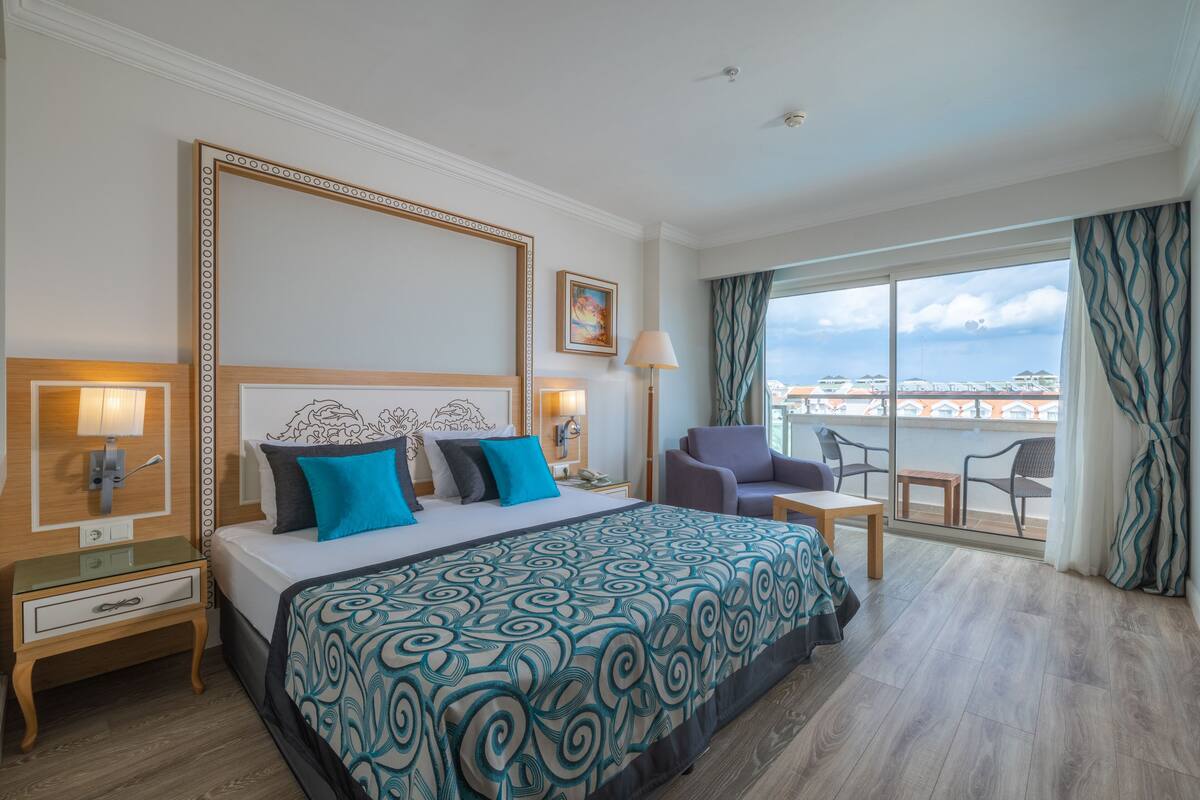 A hotel room with a large bed, a desk, a chair, and a balcony with a view of the city.