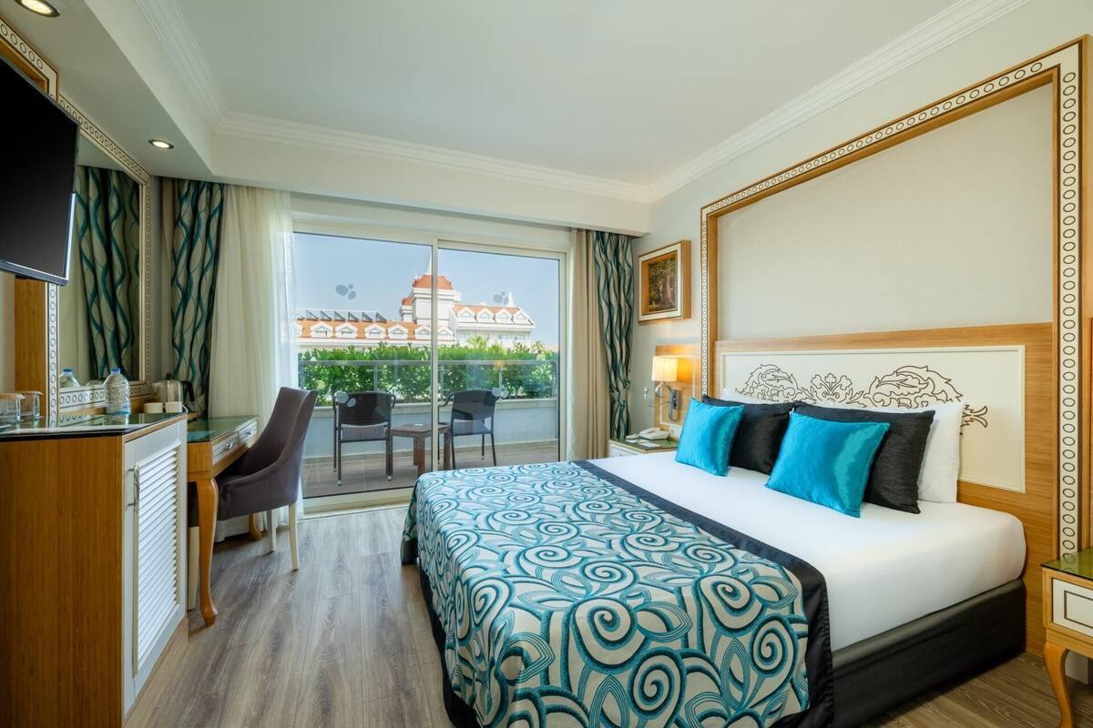 A hotel room with a bed, a desk, a chair, a TV, and a balcony with a view of buildings.