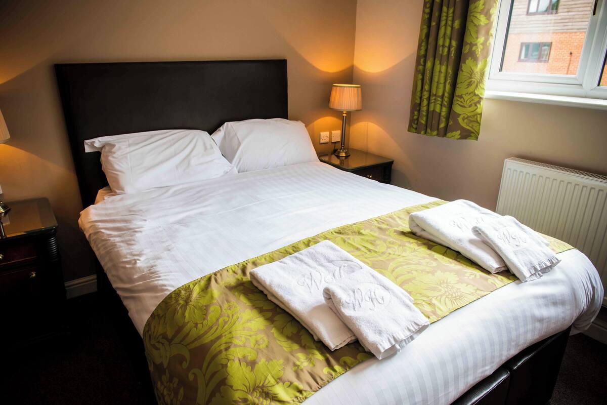 A neatly made bed with white linens and a green floral bedspread, a bedside lamp, and a window with curtains.