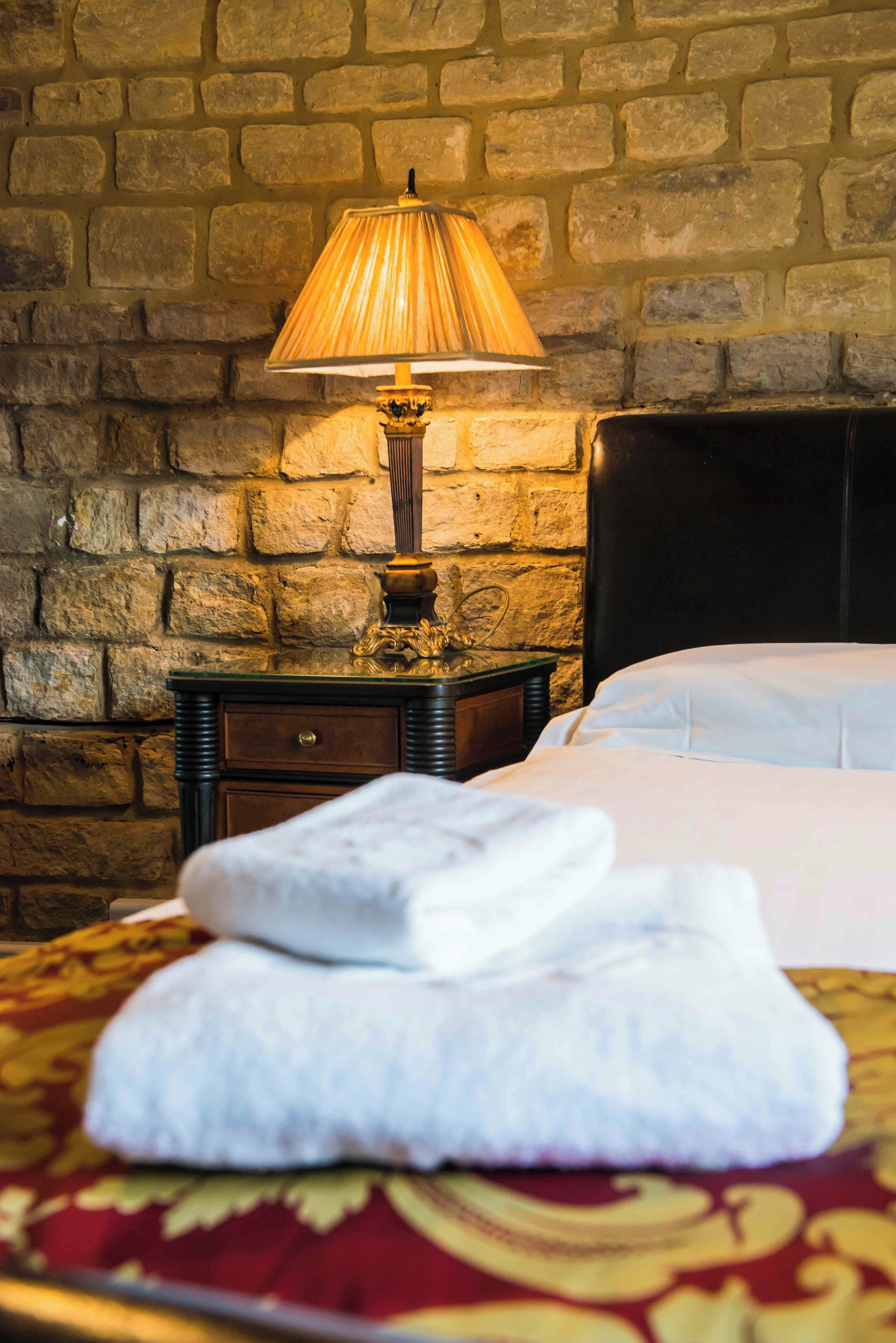 A neatly made bed with white linens, a lamp on a wooden nightstand, and a stone wall in the background.