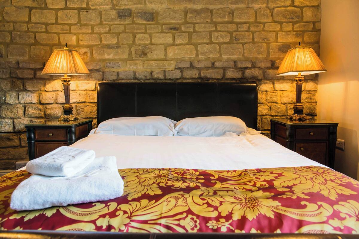A hotel room with a stone wall, a large bed with white linens, two bedside tables with lamps, and a red and gold patterned bedspread.