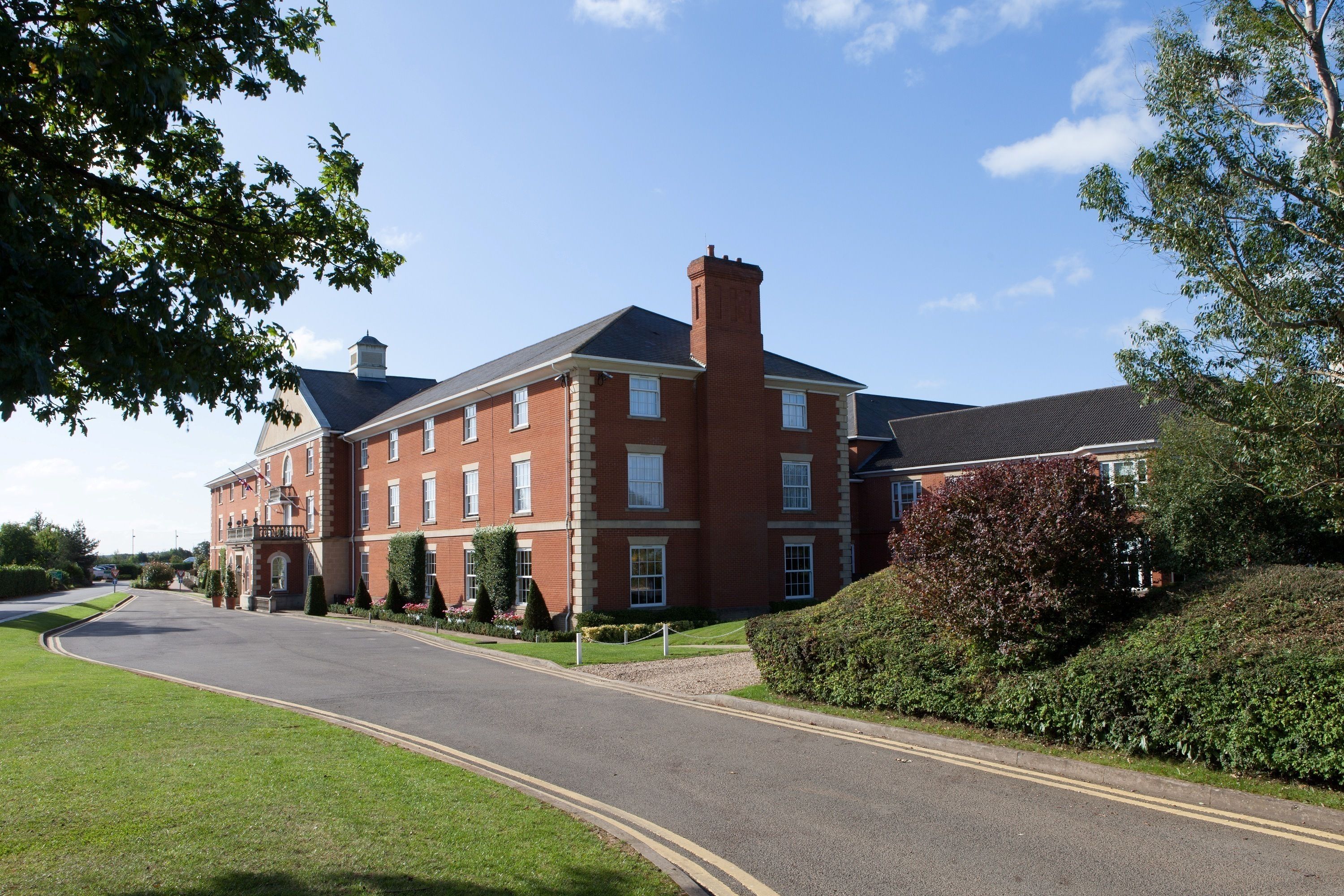 A large brick building with multiple windows and a central chimney, surrounded by well-maintained lawns and hedges.