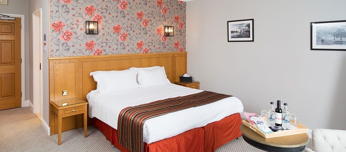 Habitación de hotel con cabecera de madera, cama grande con ropa blanca, cubrecama rojo con falda y mesita de noche con lámpara, además de bandeja con botellas y vasos.