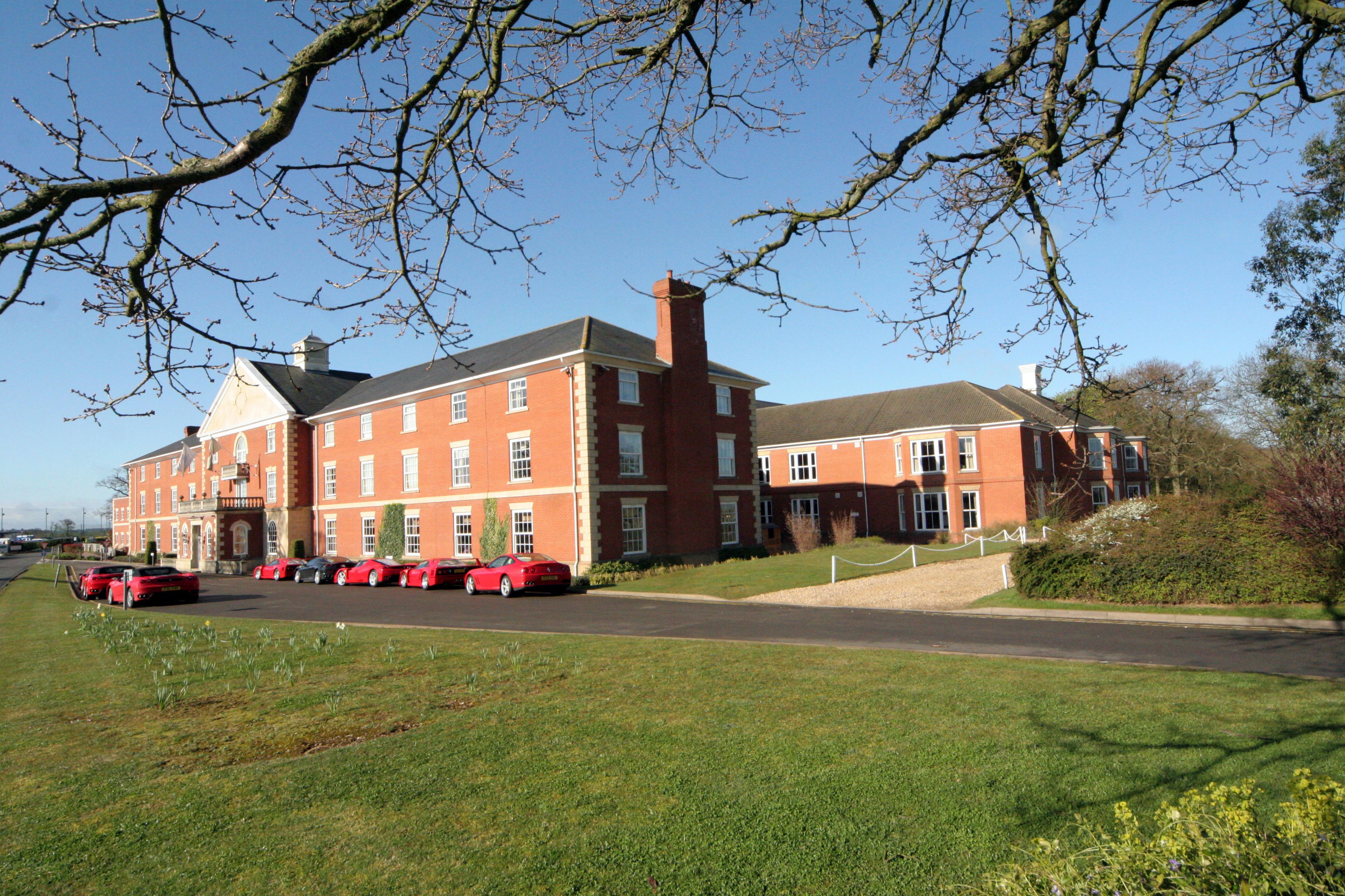 A large red brick building with multiple windows and a central chimney. There are several red cars parked in front.