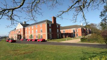 A large red brick building with multiple windows and a central chimney. There are several red cars parked in front.