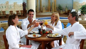 A group of people toasting at a round table with breakfast items and drinks, in a room with a pool and classical architecture. - Whittlebury Hall Hotel & Spa (Towcester)