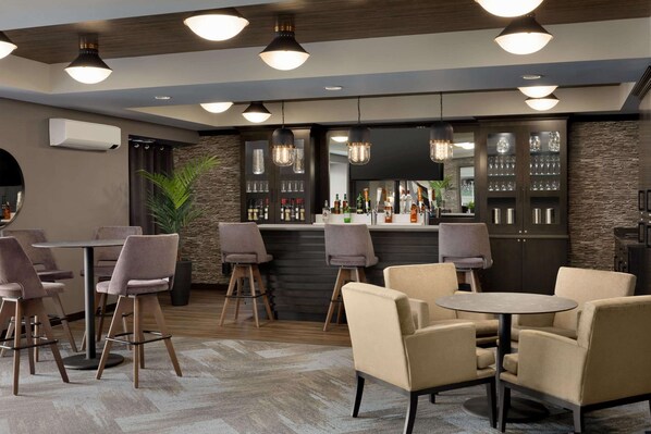 A modern bar area with round tables and bar stools, a stone accent wall, and a variety of bottles and glasses on shelves.