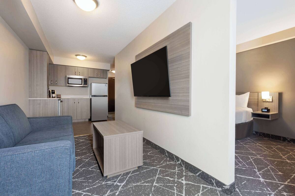 Une chambre d’hôtel moderne avec un canapé, un téléviseur à écran plat fixé au mur, une petite table de chevet avec une lampe et, au fond, une kitchenette.