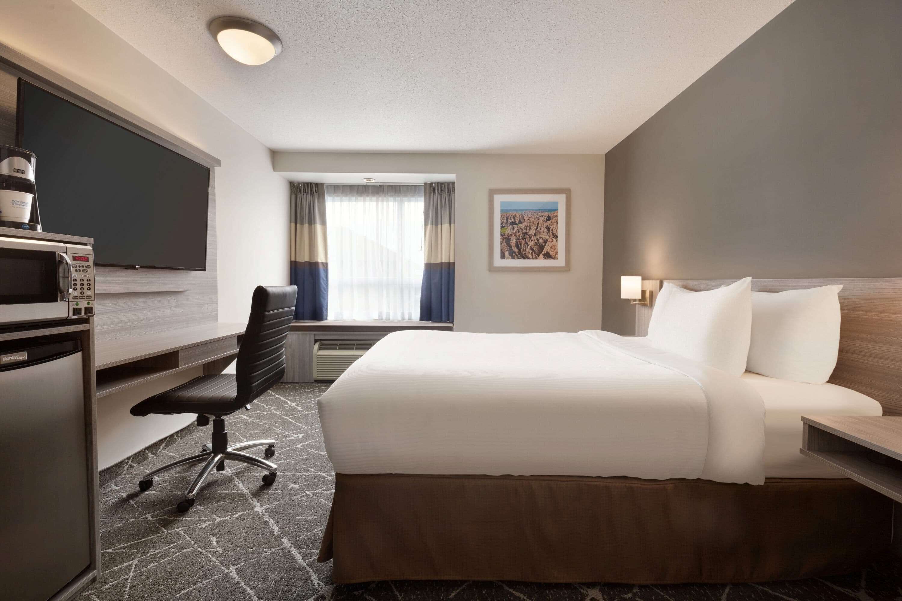 A hotel room with a bed, a desk with a computer, a TV, a microwave, a chair, and a window with curtains.