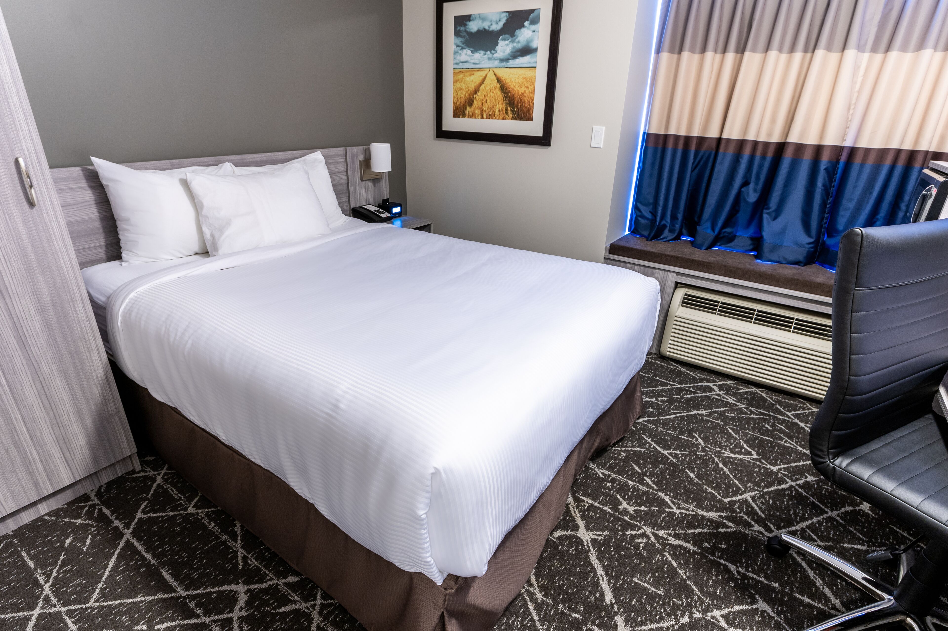 Habitación de hotel con una cama grande, una mesita de noche con lámpara, una silla, una ventana con cortinas e imagen enmarcada en la pared.