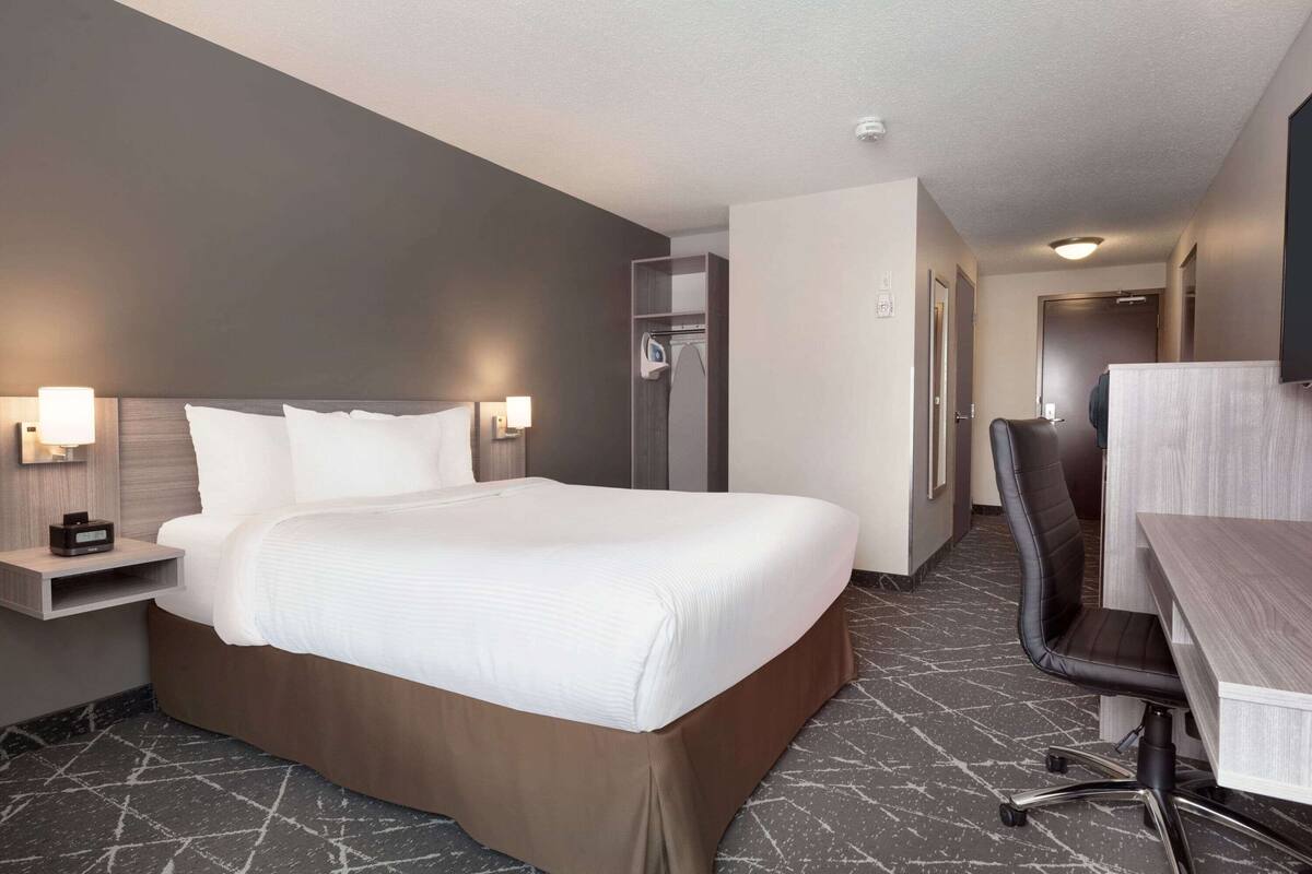 Une chambre d’hôtel avec un grand lit, un bureau et une chaise. On y trouve également une armoire et un miroir au mur.