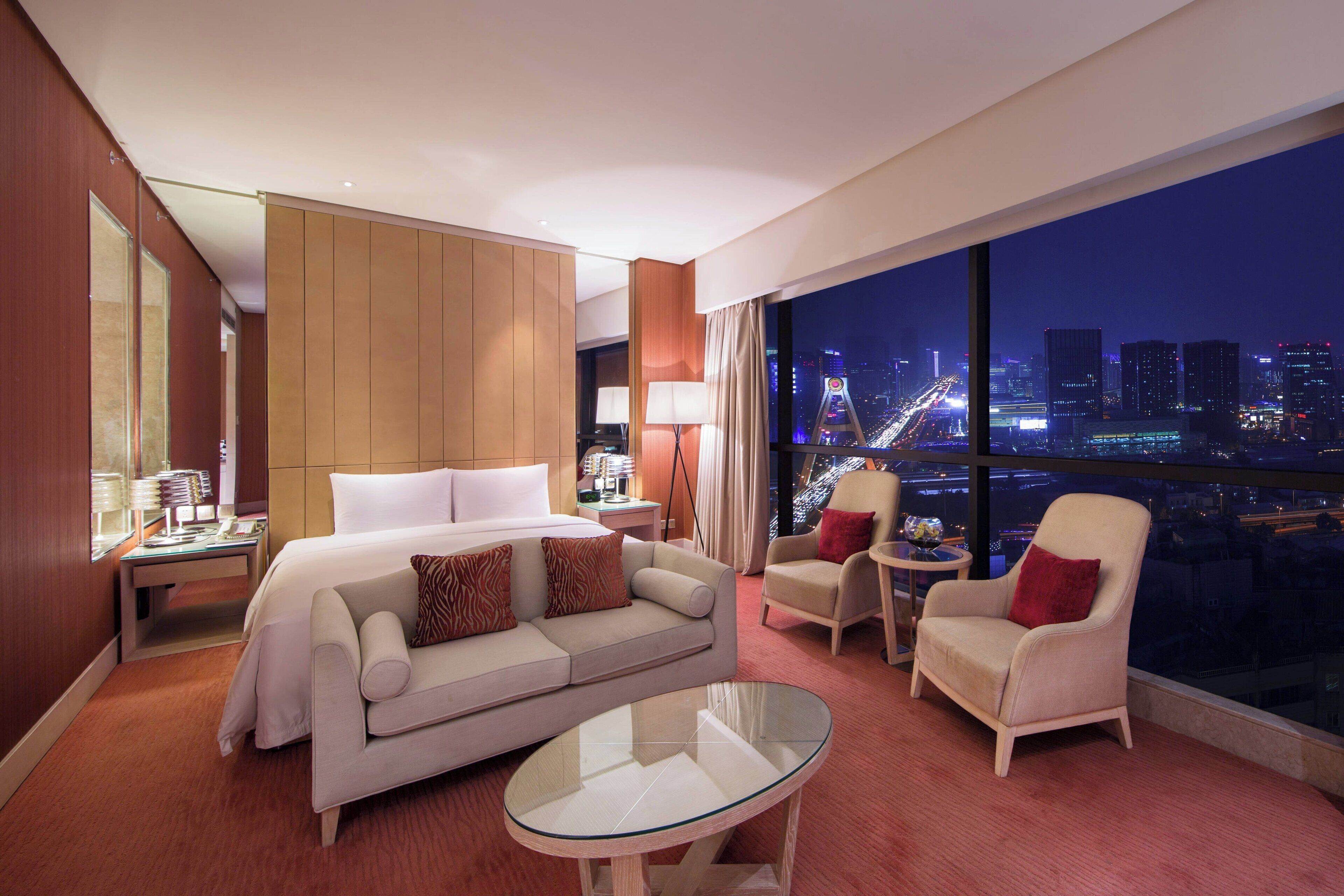 Una habitación de hotel moderna con una cama grande, un sofá, una mesa de centro de vidrio y vistas a la ciudad de noche.