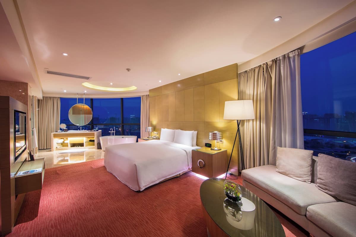 A modern hotel room with a large bed, a sofa, a glass coffee table, a desk with a lamp, and a view of the city at night.