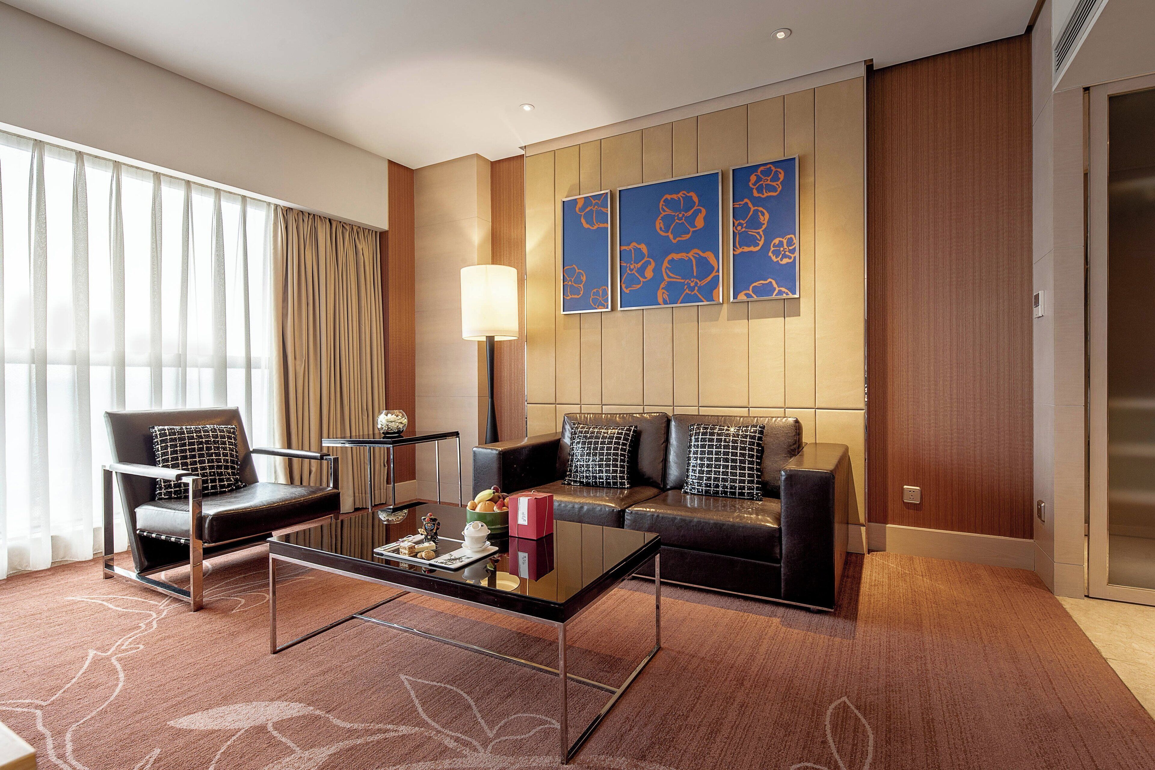 A modern hotel room with a leather sofa, a glass coffee table, a black armchair, and a large window with curtains.