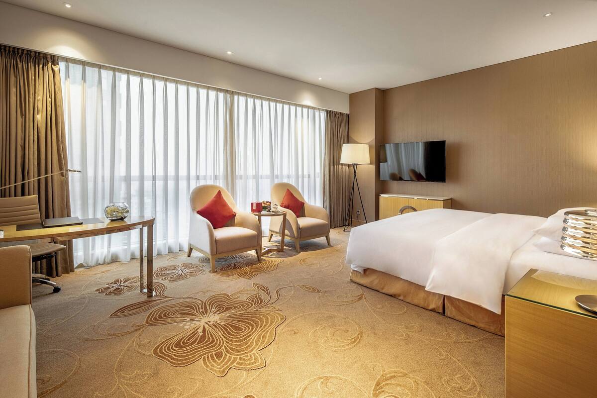 A hotel room with a bed, two armchairs, a desk, a TV, and a large window with curtains.