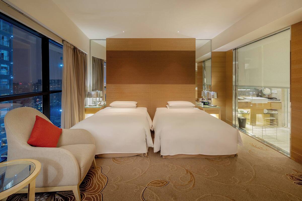 A hotel room with two beds, a sitting area with a red pillow, a glass table, and a view of the city at night.