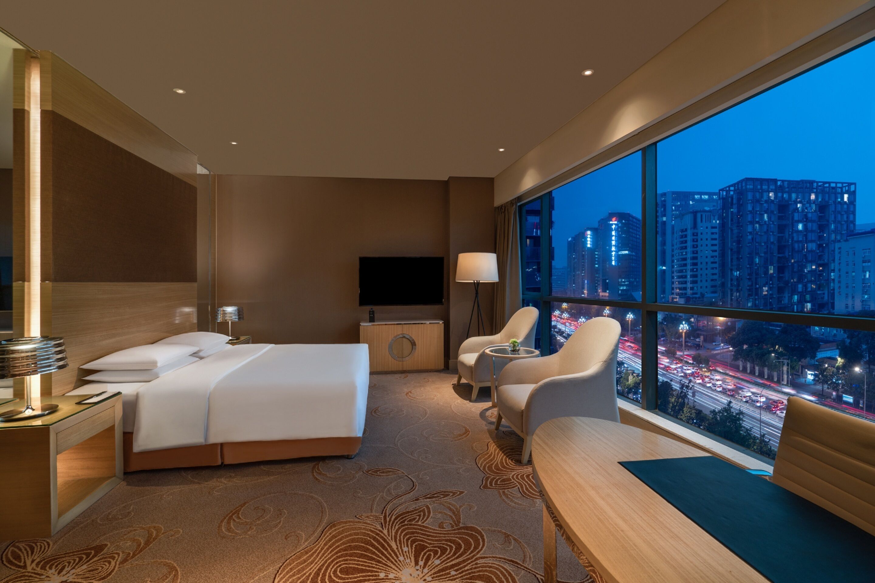 Habitación de hotel con una cama grande, televisión, escritorio, silla y vista a la ciudad de noche.
