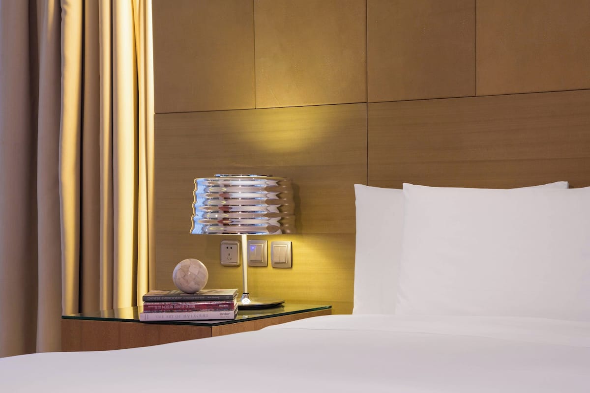 A neatly made bed with white linens, a bedside lamp, and a stack of books on a glass table beside the bed.
