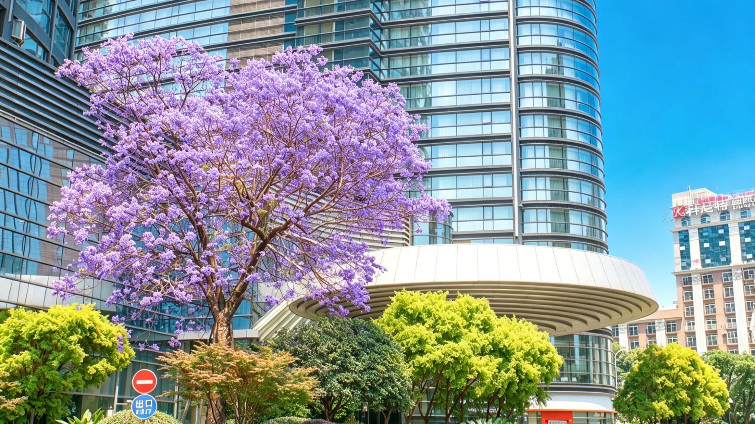 A modern building with a curved facade, a large tree with purple flowers, and a clear blue sky.