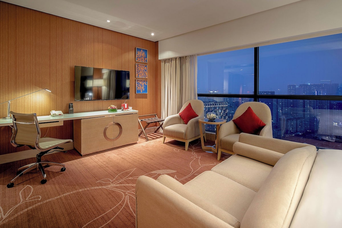 A hotel room with a large window, a desk with a chair, a sofa, and a small table with a lamp.