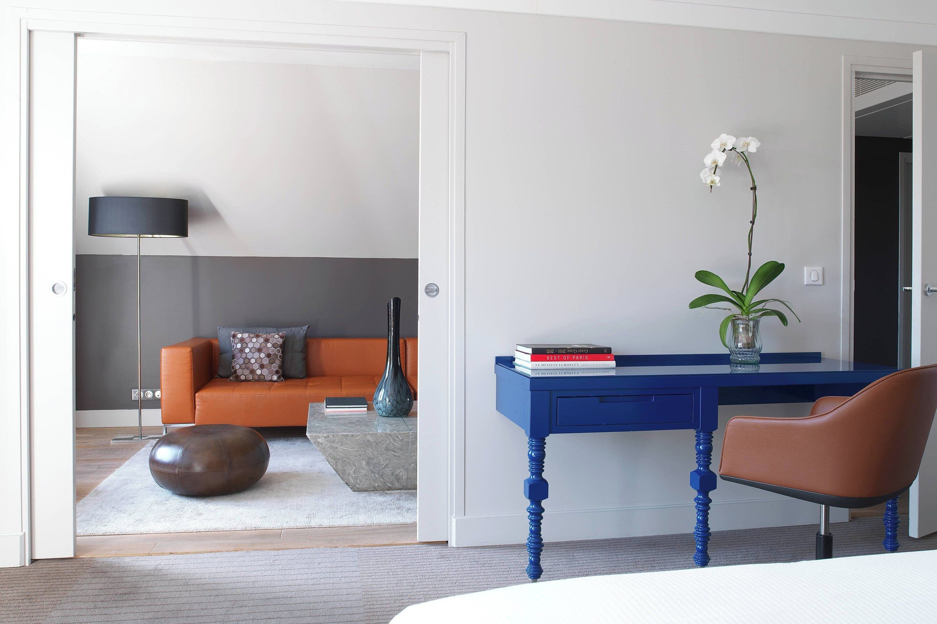 Quarto moderno com escrivaninha azul, cadeira marrom, banquinho redondo, sofá com almofadas estampadas e um vaso com orquídea.