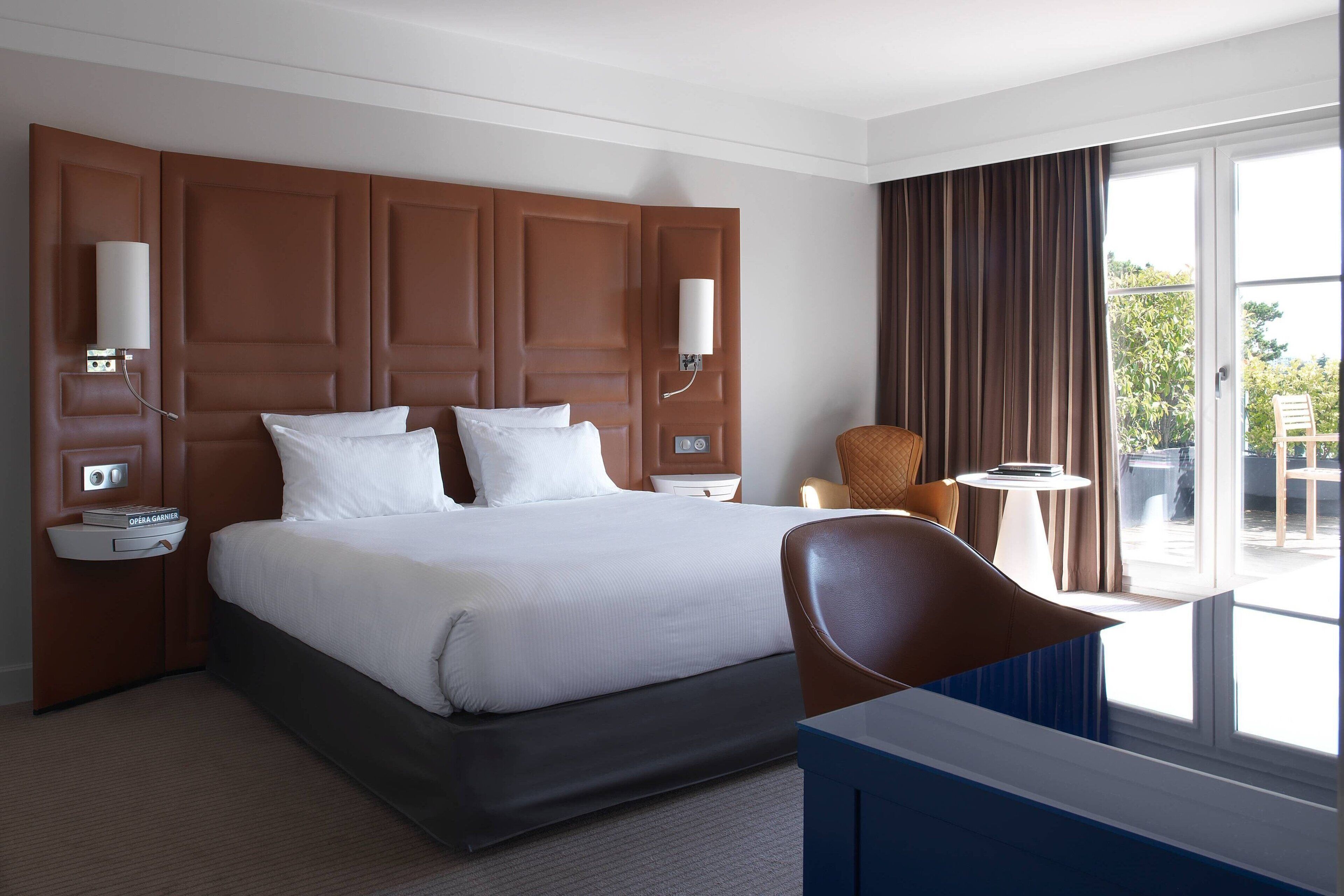 A hotel room with a large bed, a chair, a small table, and a window with curtains.