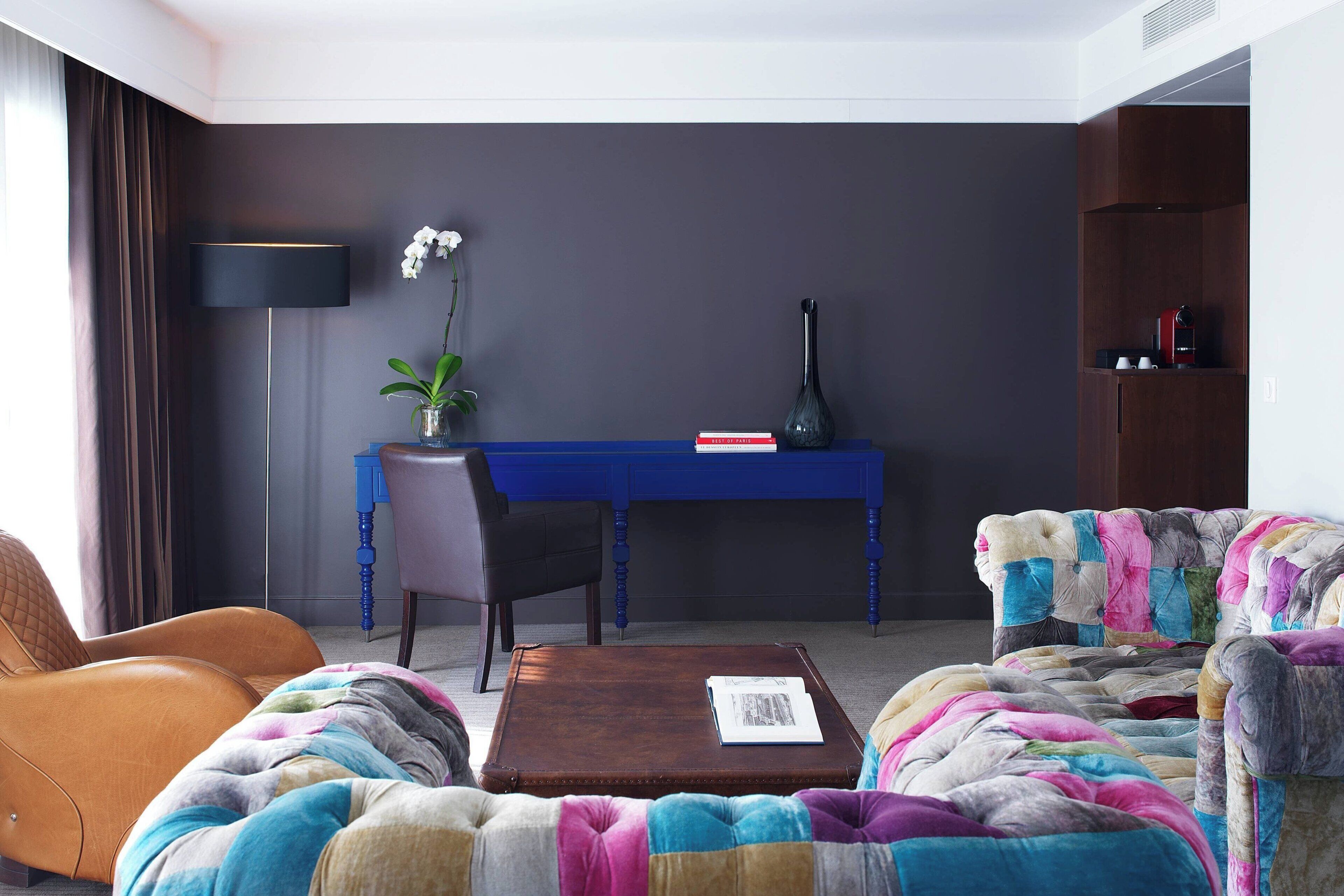 Un salon moderne avec un bureau bleu, un fauteuil en cuir marron, un canapé coloré et une table basse.
