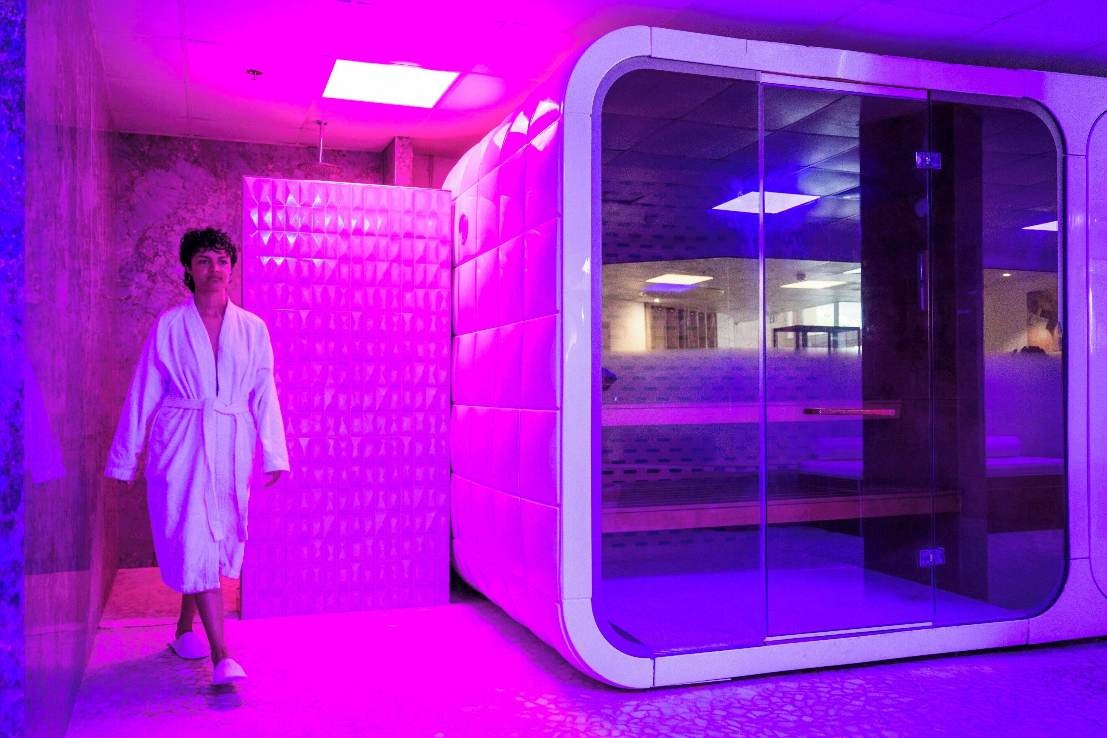 A person in a white bathrobe walking past a modern, glass-walled sauna with a tiled floor.