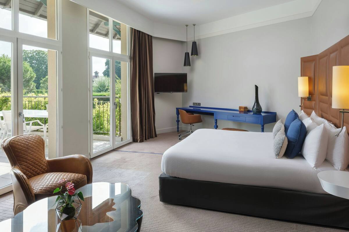 A hotel room with a large bed, a desk, a chair, a TV, and a view of the outdoors.