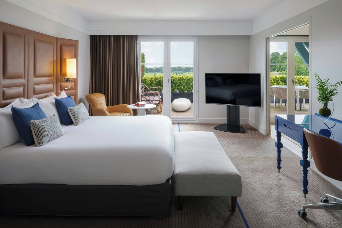 A hotel room with a large bed, a TV, a balcony with outdoor furniture, and a view of greenery.