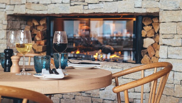 A stone fireplace with a fire, a round wooden table with chairs, and wine glasses. - Renaissance Paris Hippodrome De St. Cloud Hotel (Rueil-Malmaison)