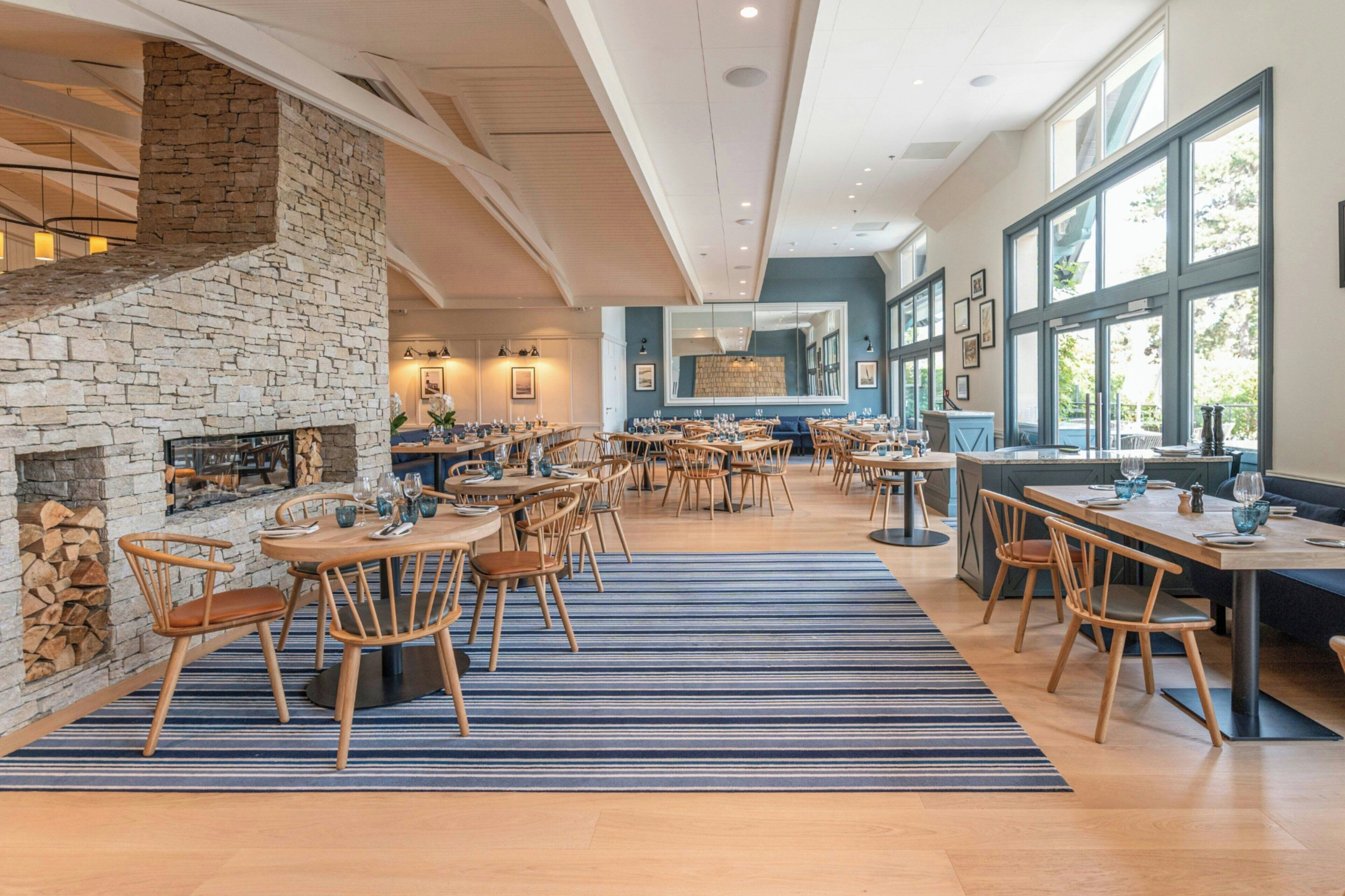 A spacious dining area with multiple tables and chairs, a stone fireplace, and large windows allowing natural light.