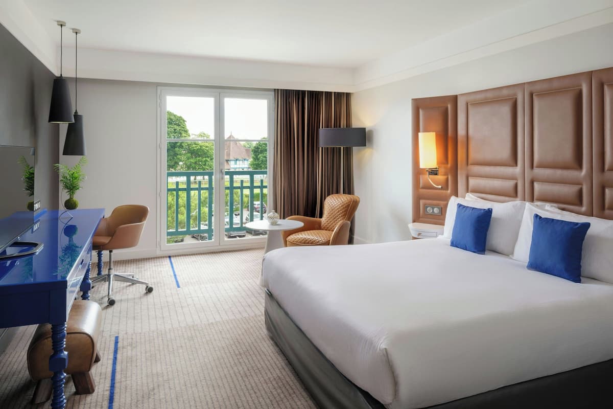 A hotel room with a large bed, a desk with a chair, a small round table, and a balcony with a view of greenery.