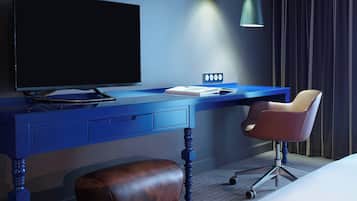 A modern hotel room with a blue desk, a television, a chair, and a bedside table.